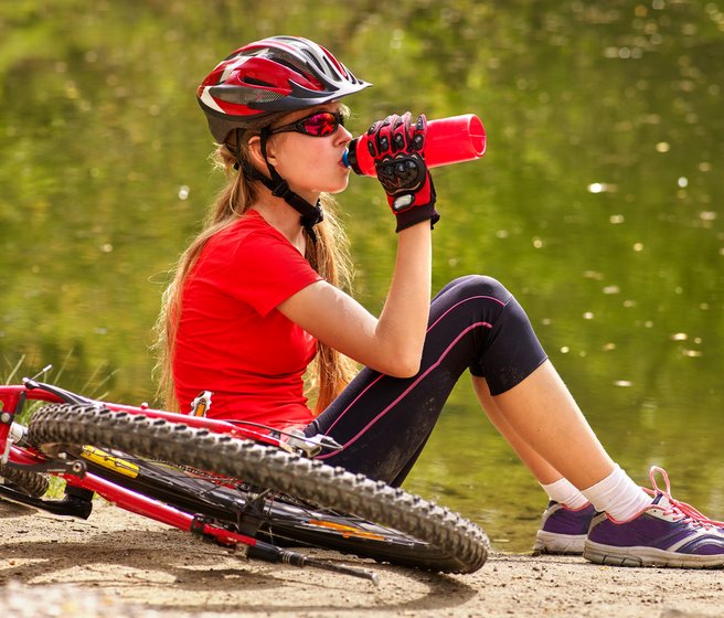 Mädchen in Sportkleidern sitzt neben einem Mountainbike und trinkt aus einer Flasche
