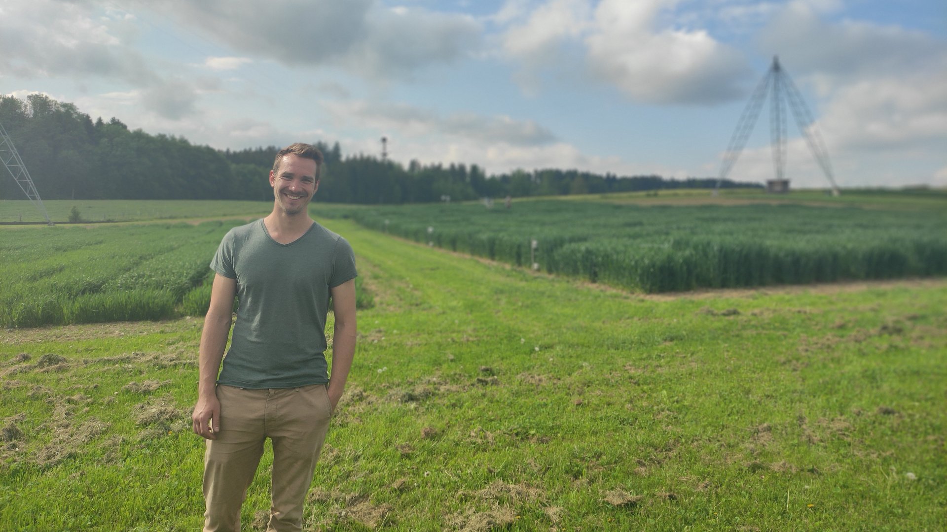 Junger Forscher vor einem grünen Feld, im Hintergrund eine grosse dreibeinige Stahlkonstruktion