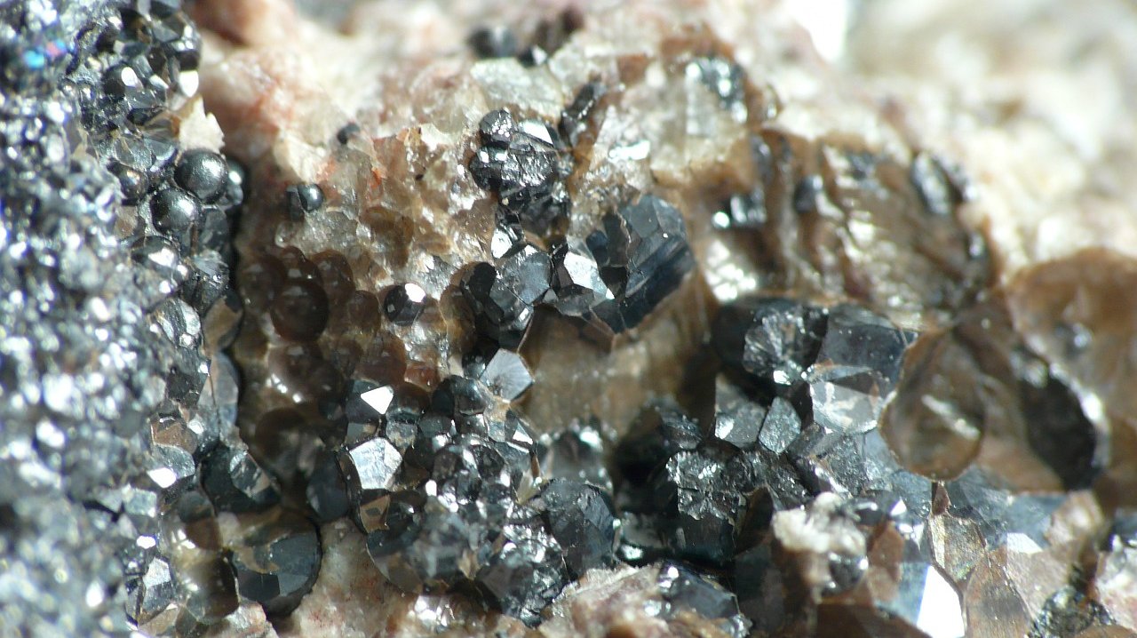 Dunkle Kristalle und kugelige Aggregate des Minerals Pechblende auf einem Untergrund von hellerem Kristall