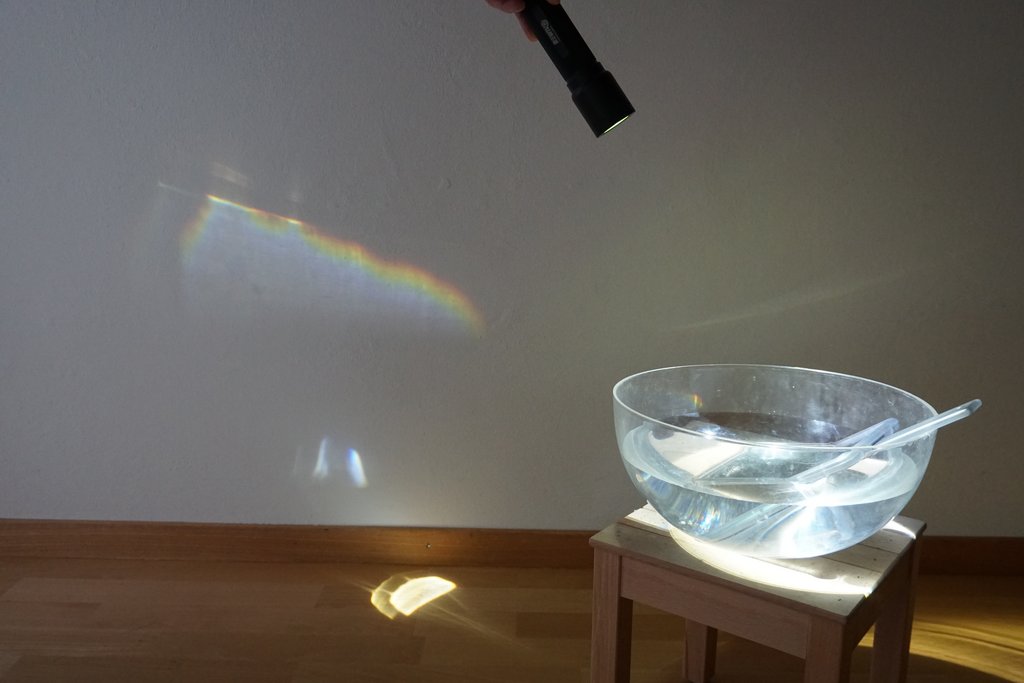 Schüssel mit Wasser und Spiegel in einem abgedunkelten Raum, Regenbogenprojektion an der Wand