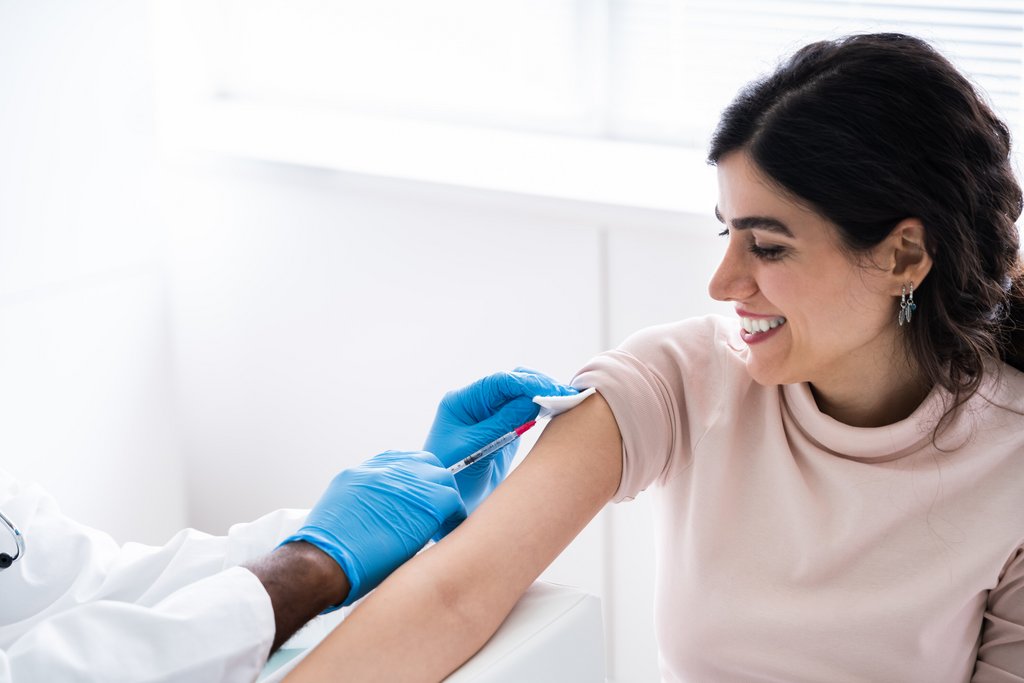 Frau mit hochgerolltem Ärmel schaut zu, wie die Hand eines Arztes mit Gummihandschuh die Impfnadel am Oberarm ansetzt