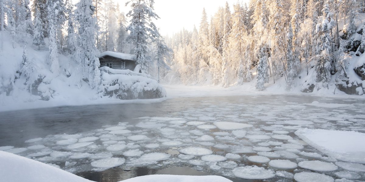 Petit lac en partie gelé dans une forêt enneigée, avec une cabane à l’arrière-plan.