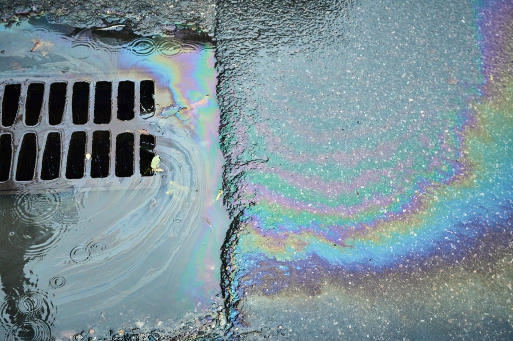 Farbig schillernde &Ouml;lschlieren auf dem feuchten Asphalt neben einem Gully